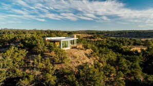 Ett modernt hus med ett platt tak, stora fönster och en terrass, omgivet av tät skog och kullar.
