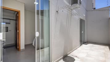 Un espacio residencial moderno con un cerramiento de vidrio que da a un baño, un patio con baldosas y una pared exterior blanca.