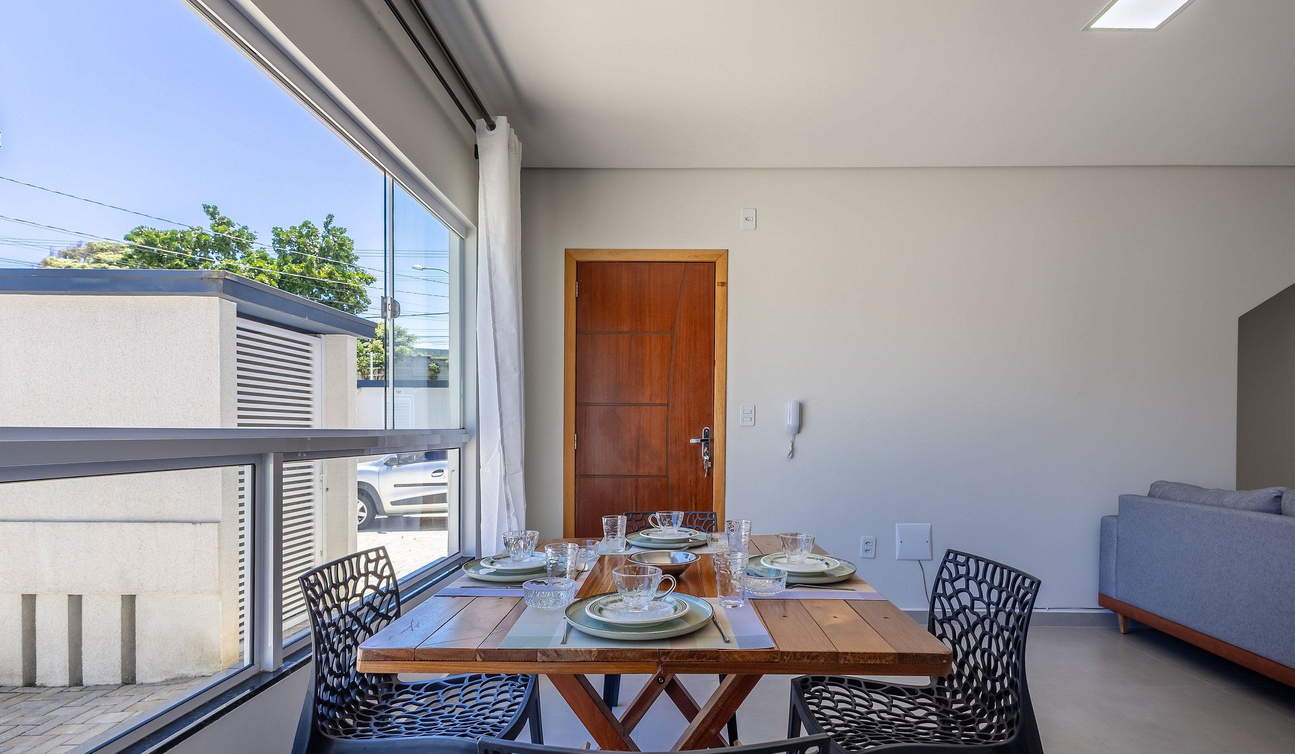 A dining area with a wooden table set for four, surrounded by black chairs, a grey sofa, and a wooden door.