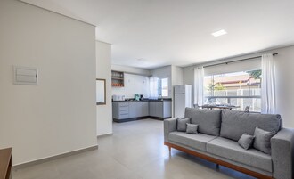 Uma sala de estar moderna com um sofá cinzento, um espaço de jantar com mesa e cadeiras, e uma cozinha ao fundo.