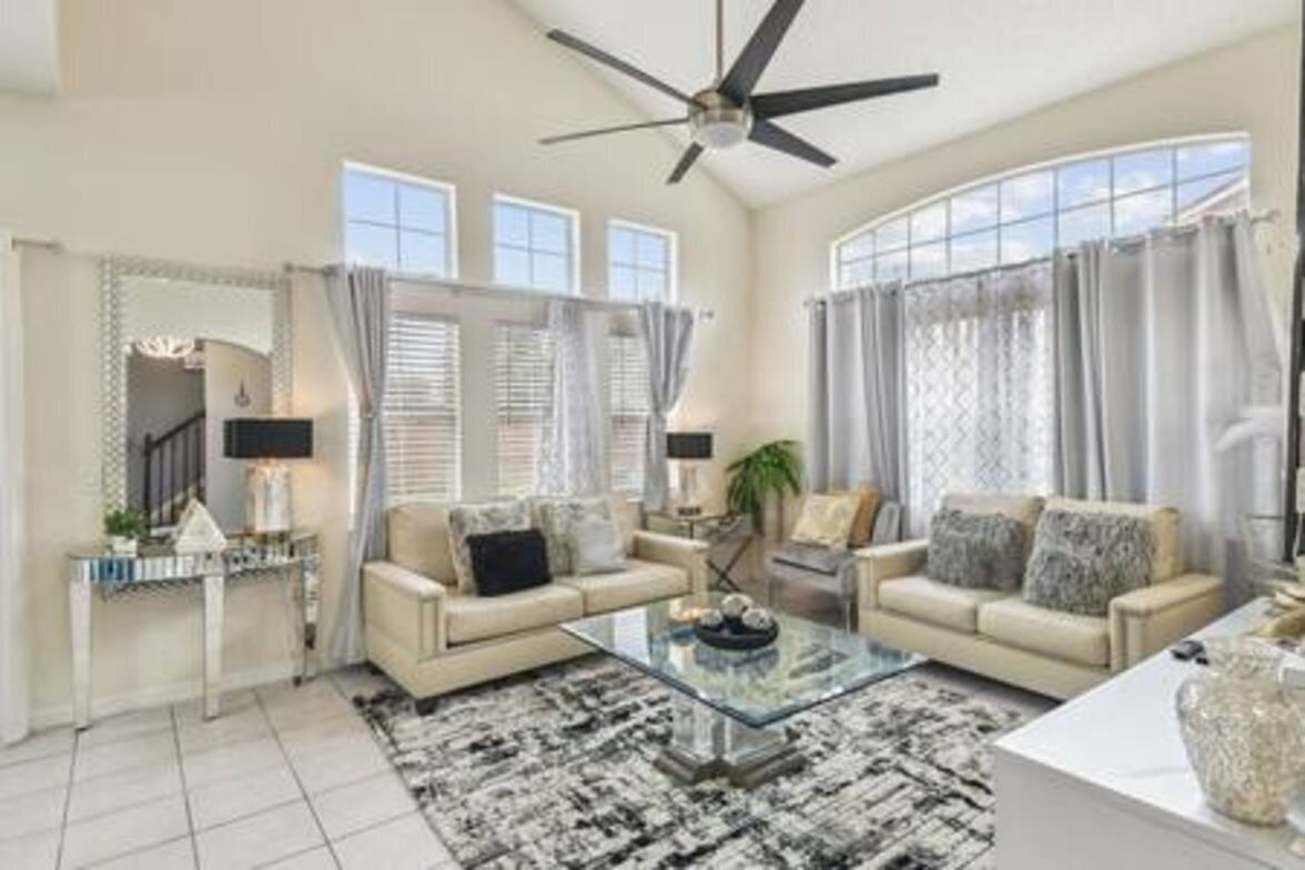 A spacious living room with a high ceiling fan, large windows with curtains, a beige sofa set, a glass coffee table, and a modern console table.