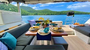 Un pont de bateau avec une table dressée pour un repas, un coin salon confortable et une vue sur un lac, avec des collines en arrière-plan.