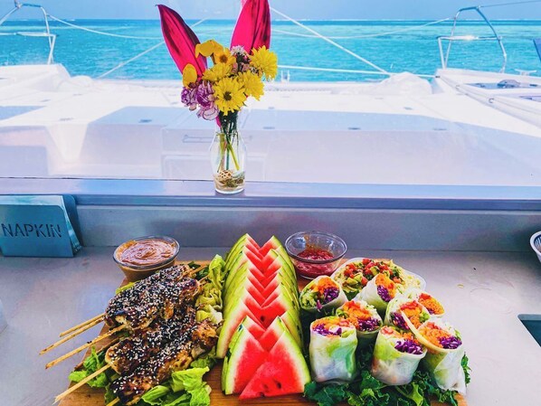 Une table dressée avec divers plats, notamment des brochettes, des tranches de pastèque et des rouleaux de printemps, sur un bateau d’où l’on peut admirer l’océan.