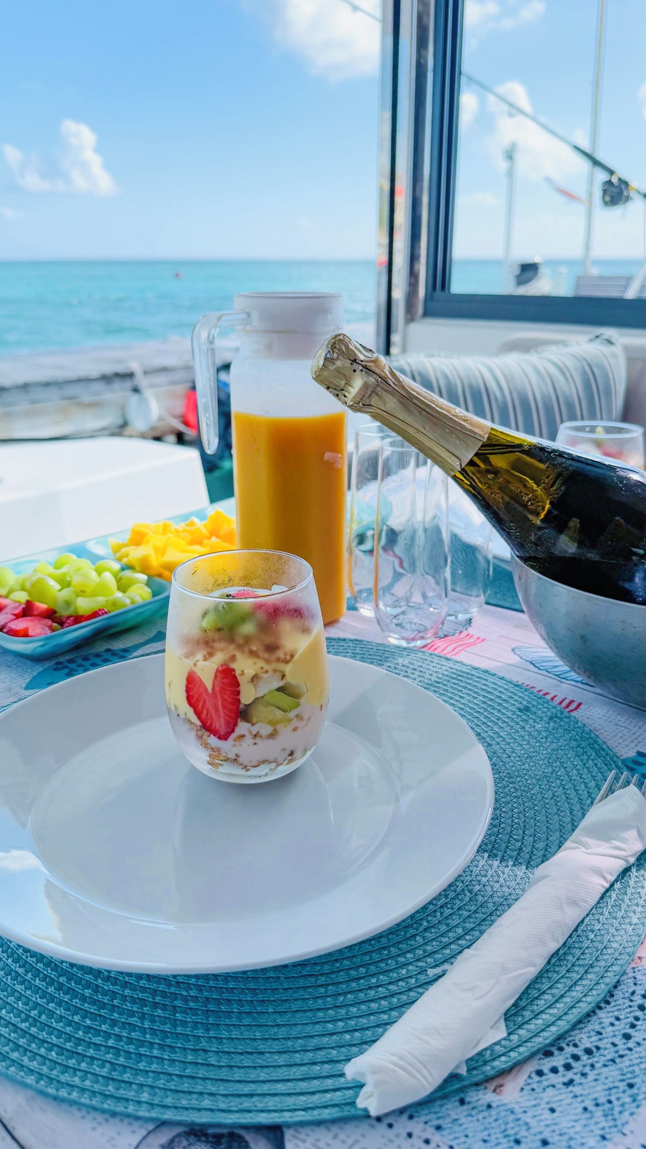 Ein Dessert auf einem Teller, eine Champagnerflasche, eine Karaffe Orangensaft und eine Fruchtschale auf einem Tisch vor einem Fenster mit Meerblick.