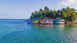 Une rangée de bungalows colorés, construits sur pilotis au-dessus de l’eau, avec une eau d’un bleu cristallin et des palmiers en arrière-plan.