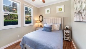 A bedroom with a bed, a ceiling fan, a nightstand with a lamp, and a window with a view of greenery.