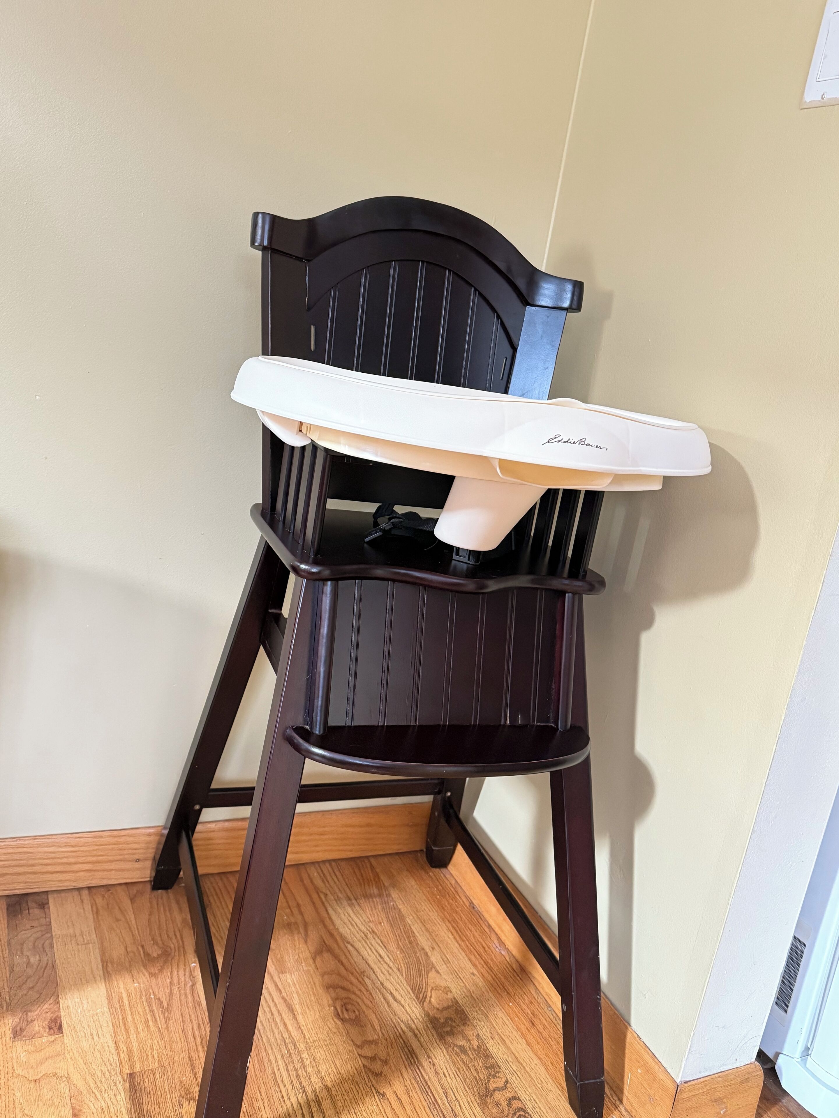 A dark wooden high chair with a white tray, positioned against a wall with a wooden floor.