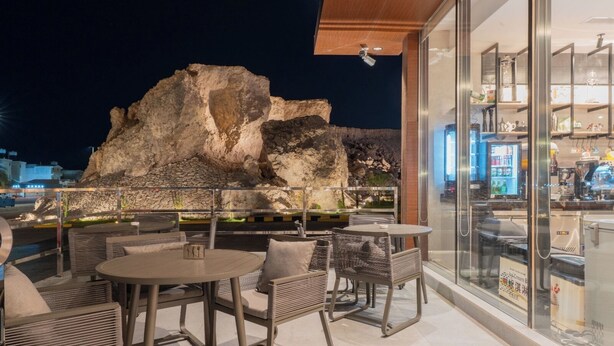 Un área de estar al aire libre con mesas y sillas, una formación rocosa al fondo y una pared de vidrio que muestra un bar y una cocina en el interior.