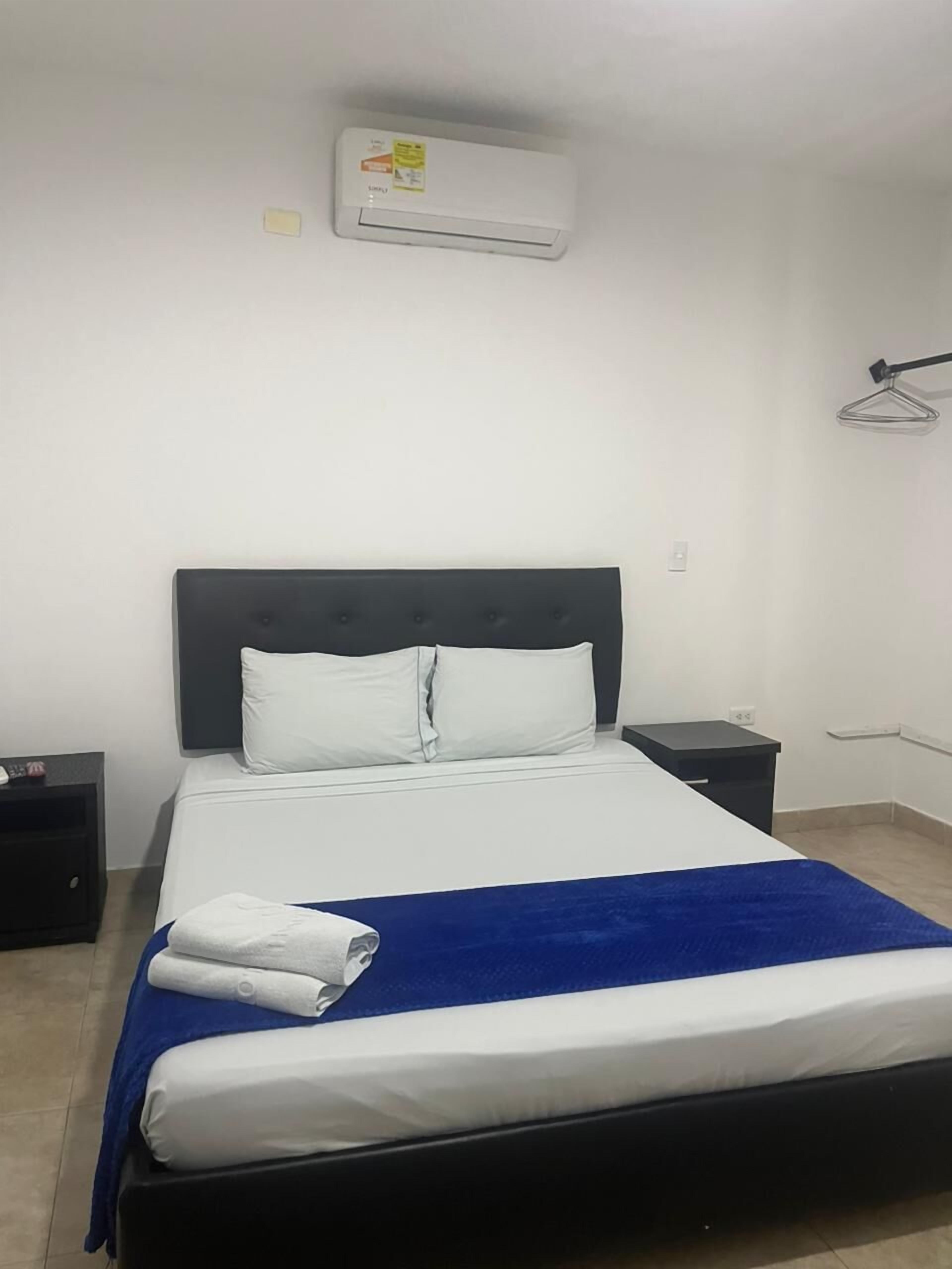 A neatly made bed with a blue blanket, white pillows, and a folded white towel. There is a black nightstand with a lamp on it, and a wall-mounted air conditioner above the bed.