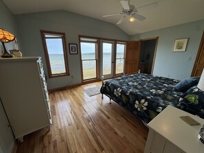 A bedroom with a bed, a dresser, a desk, a ceiling fan, and large windows with a view of the water.