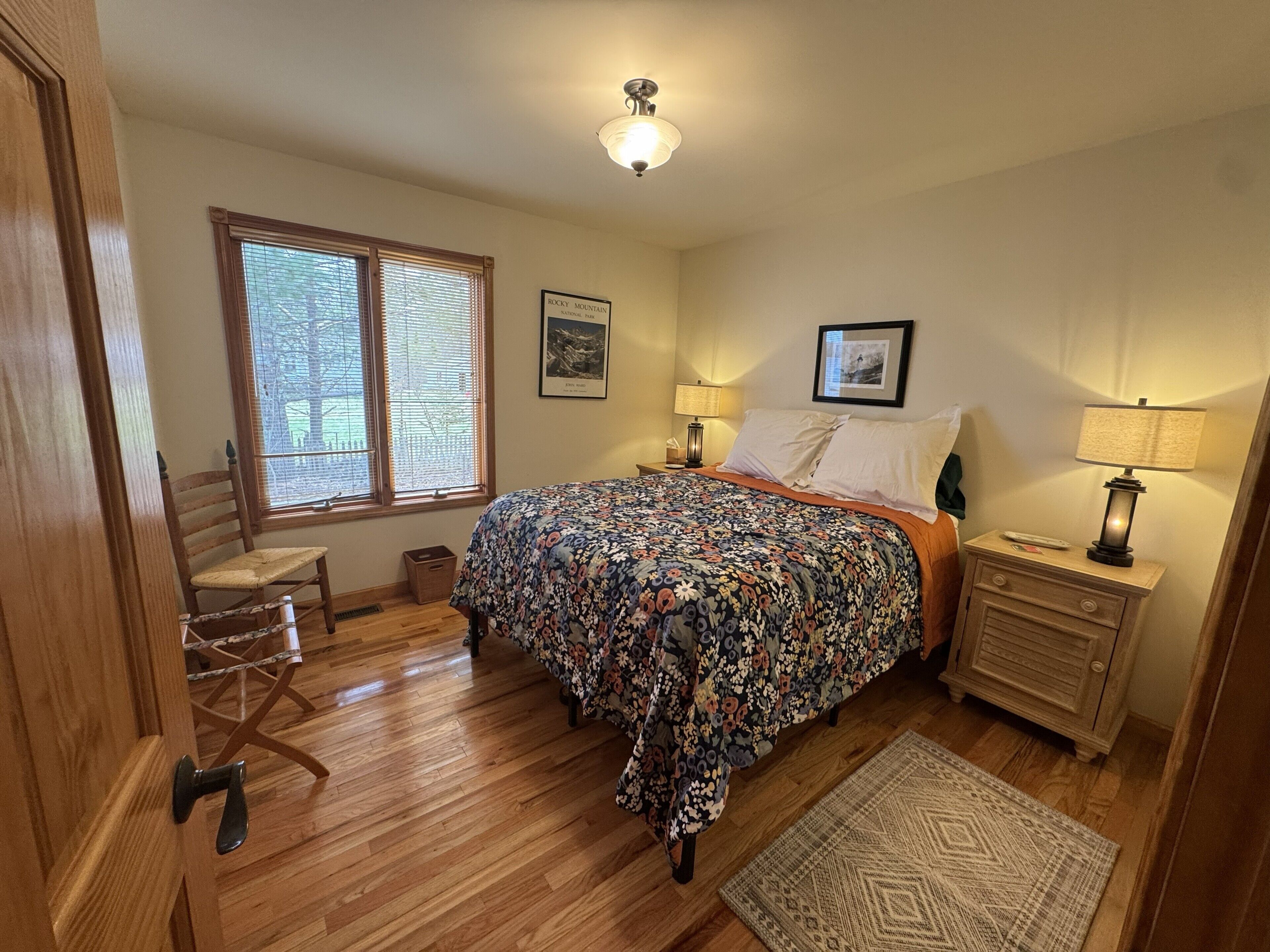 Une chambre à coucher comprenant un lit, une chaise, une table de chevet, une lampe, une fenêtre avec des stores et un luminaire au plafond.