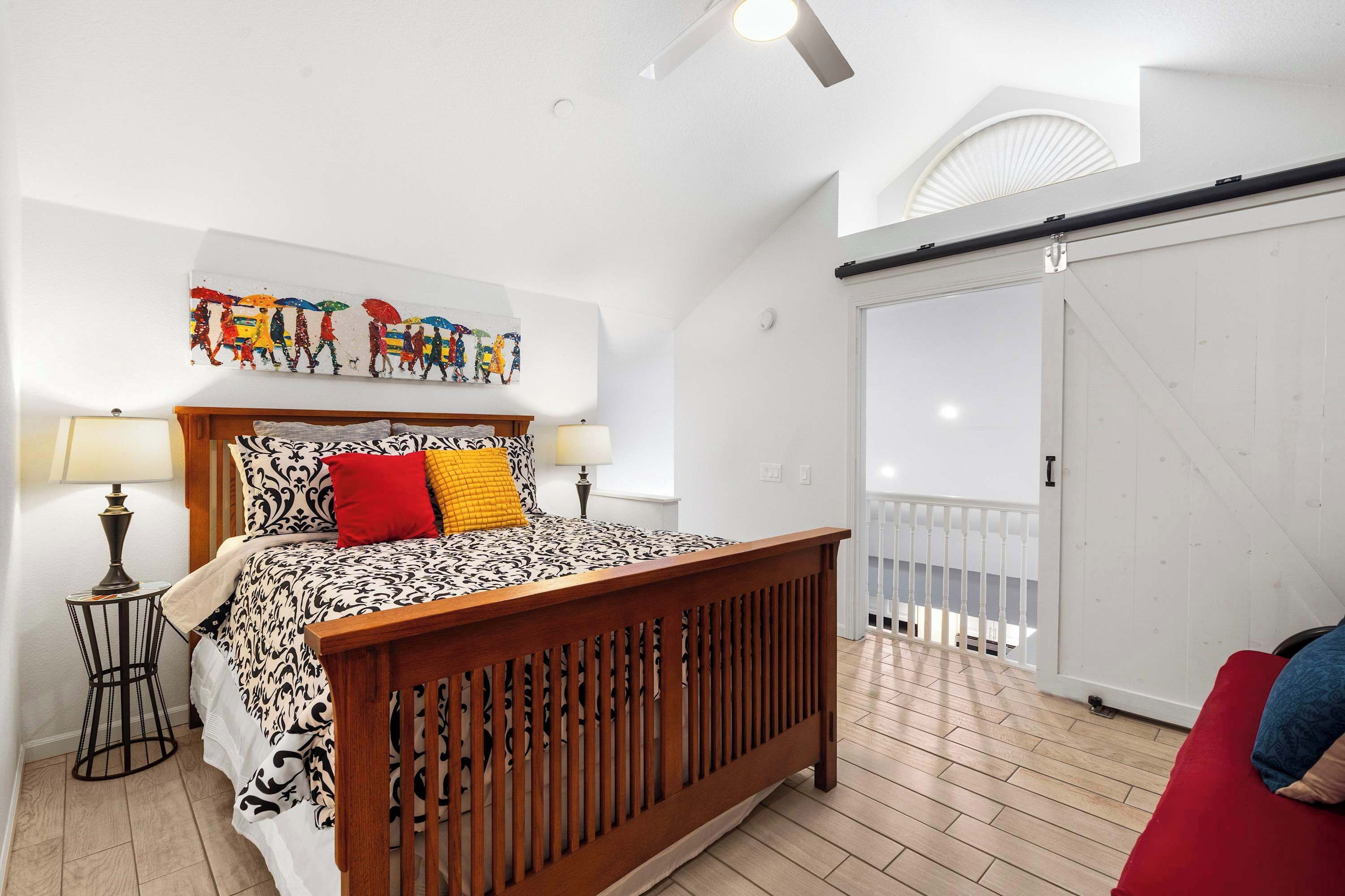 Un dormitorio con una cama de madera, una mesita de noche, una lámpara, una obra de arte colorida en la pared y vistas al balcón.