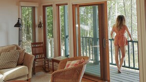 A spacious living room with a wooden ceiling, a sofa, a wicker chair, and a table. There are large sliding glass doors leading to a deck with a view of trees. - Private lakeside hideaway, 4 bedroom one-story home with stunning water views. (Sebago)
