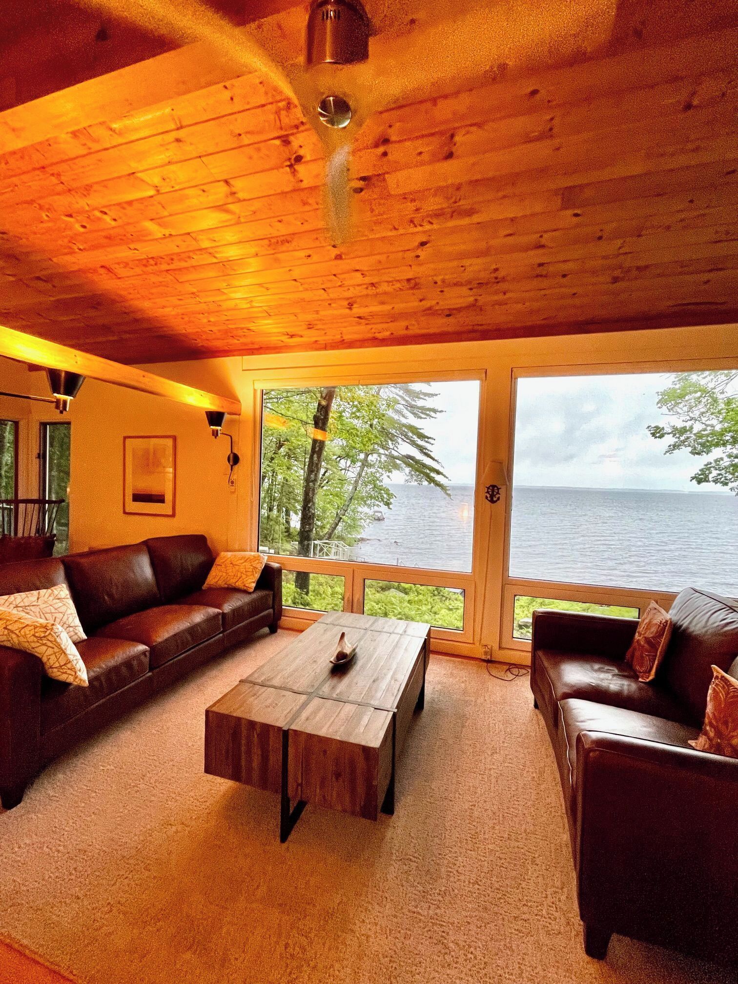 Ein gemütliches Wohnzimmer mit Holzbalkendecke, einem großen Fenster mit Blick auf ein Gewässer und einem braunen Ledersofa.