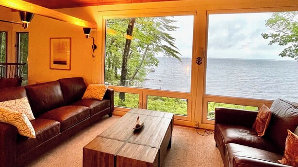 A cozy living room with a wooden ceiling, a large window overlooking a body of water, and a brown leather sofa. - Private lakeside hideaway, 4 bedroom one-story home with stunning water views. (Sebago)