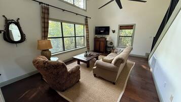 A spacious living room with a high ceiling, large windows, a ceiling fan, a fireplace, and a television.