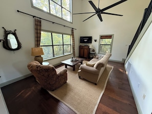 A spacious living room with a high ceiling, large windows, a ceiling fan, a fireplace, and a television.