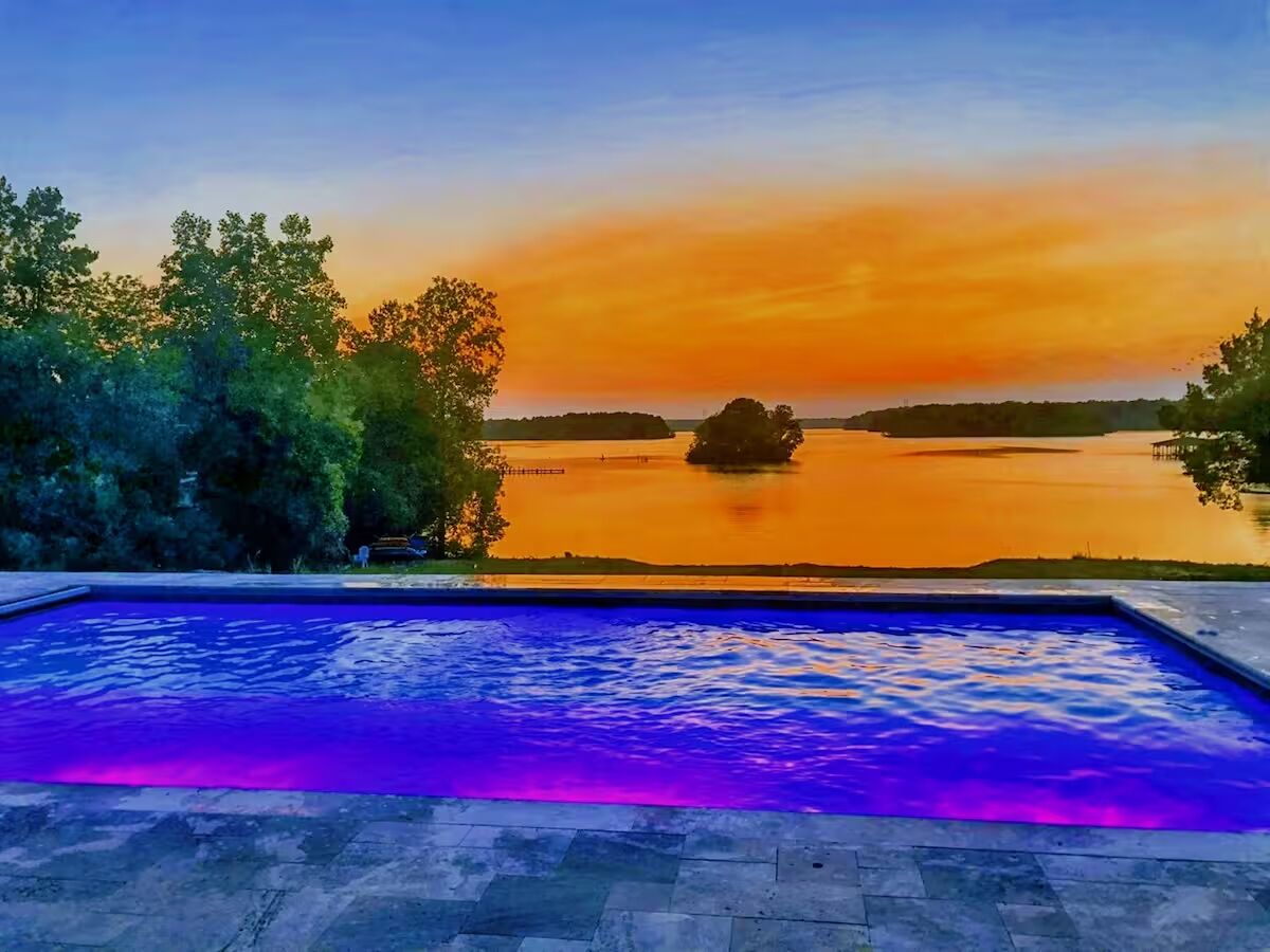 A swimming pool with a view of a sunset over a lake, surrounded by trees.