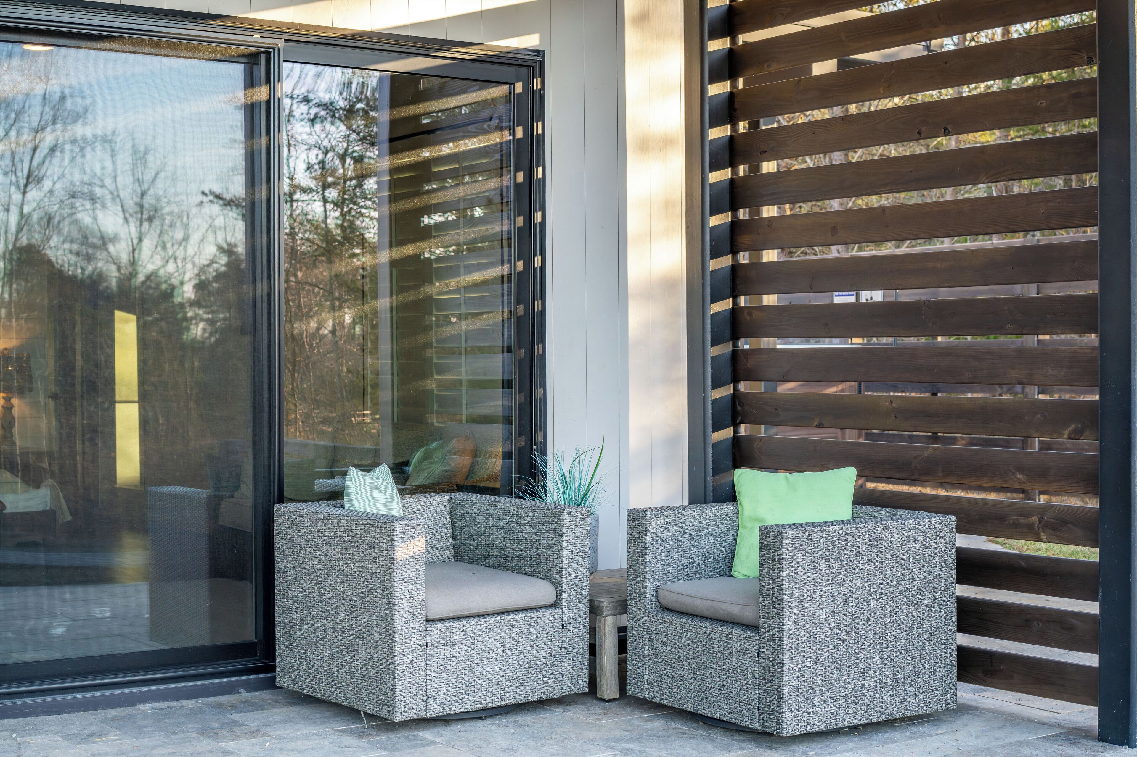 A modern outdoor seating area with two wicker chairs, one with a green cushion, and a glass door reflecting the surrounding trees.