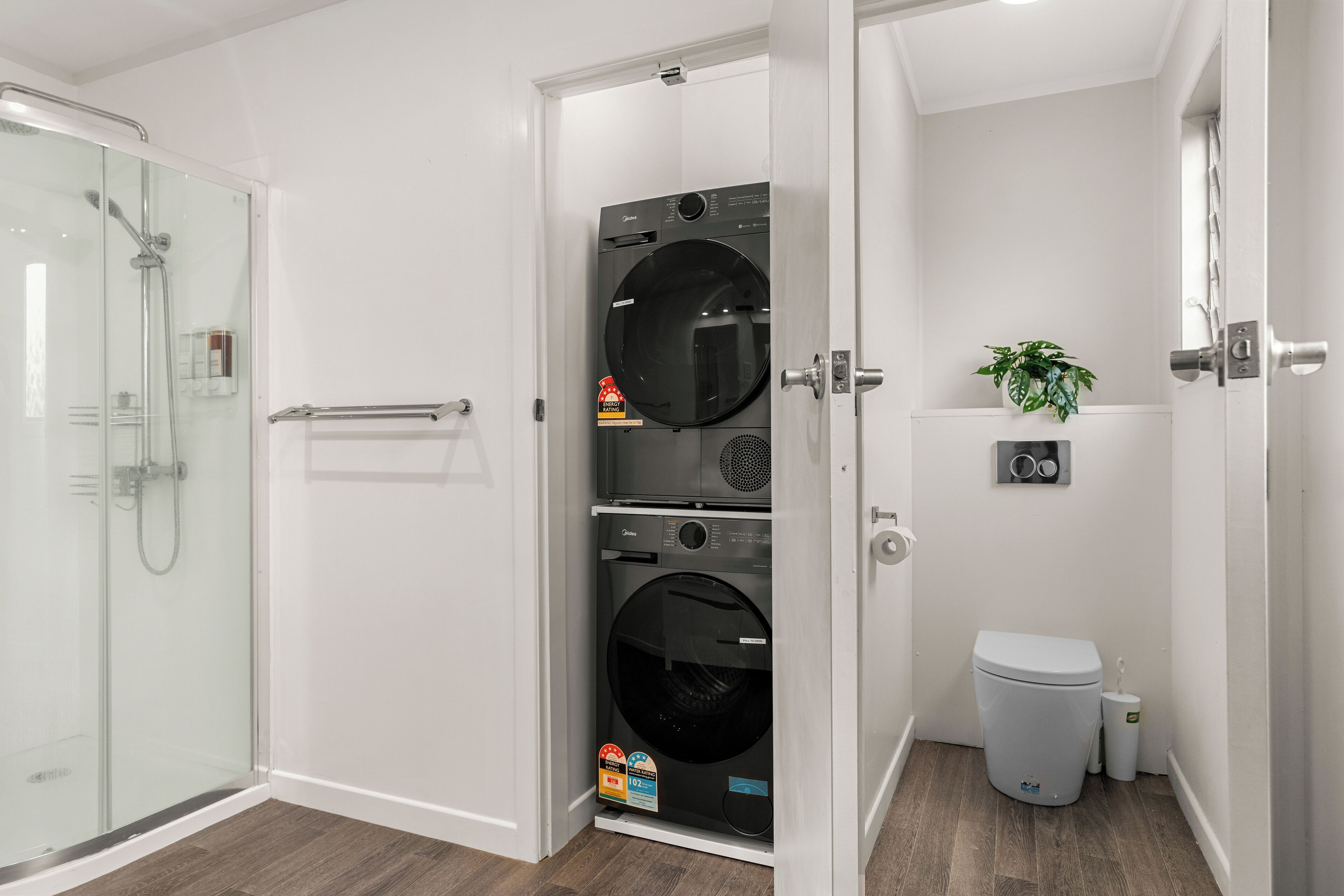 Un bagno compatto con box doccia in vetro, lavatrice e asciugatrice affiancate, un water e una piccola pianta in vaso.