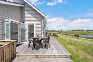 A house with a deck, a table with chairs, and a view of a river and landscape. - Sunlit Shores - Papamoa Holiday Homes (Papamoa)