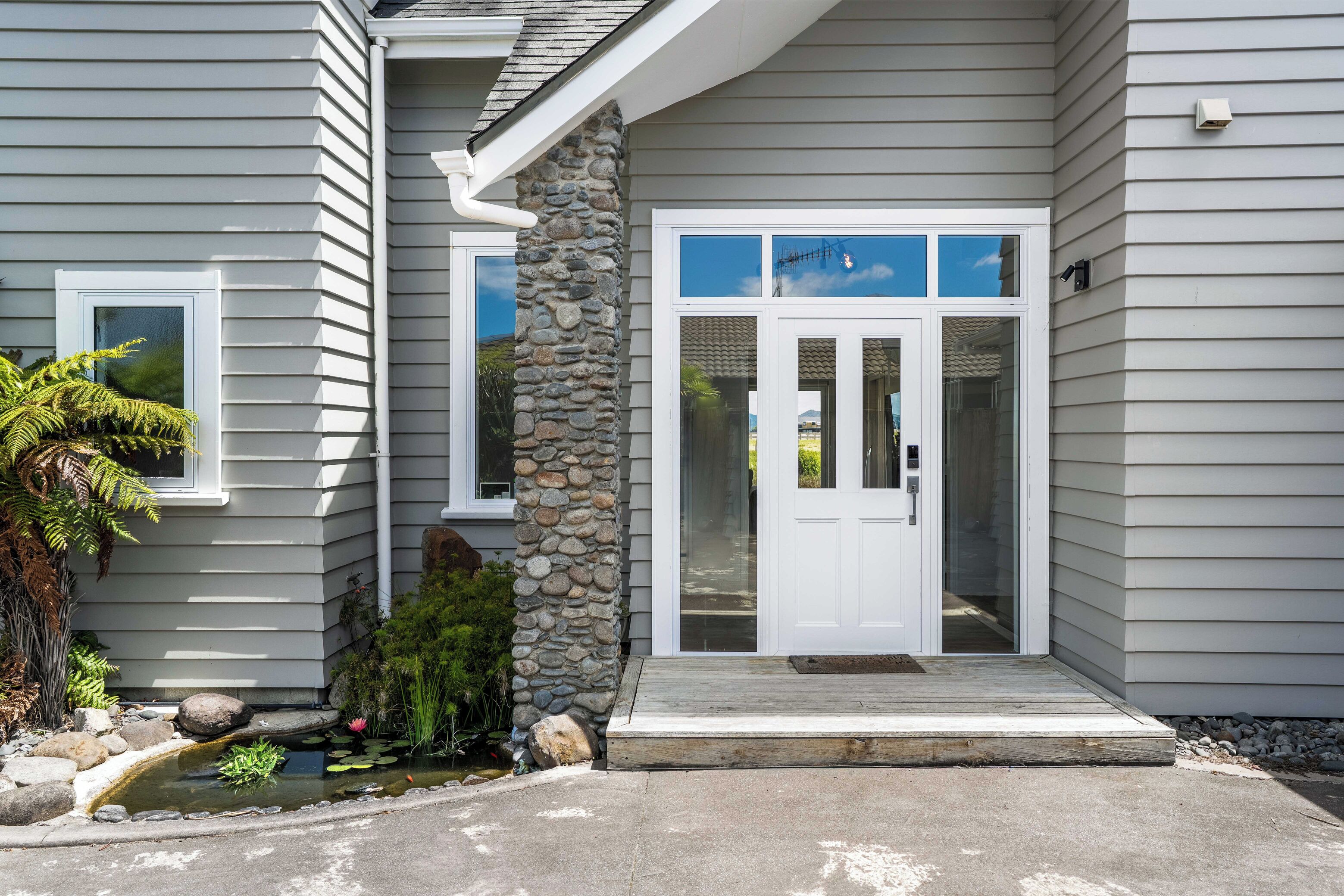 Une maison avec un pilier en pierre, une porte d’entrée avec des panneaux de verre, un petit étang avec des rochers et des plantes, et une allée en béton.