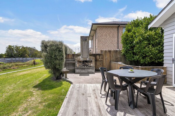 An outdoor patio with a round table and chairs, a stone fireplace, and a wooden fence. - Sunlit Shores - Papamoa Holiday Homes (Papamoa)