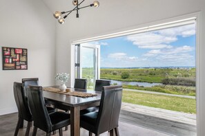 A dining area with a table and chairs, a view of a river and landscape, and a modern chandelier. - Sunlit Shores - Papamoa Holiday Homes (Papamoa)