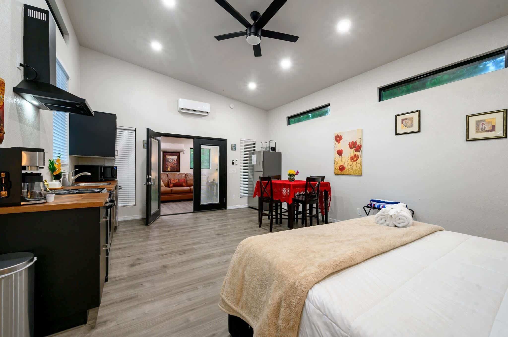 A modern hotel room with a kitchenette, a bed with a beige blanket, a dining table with a red tablecloth, and a ceiling fan.