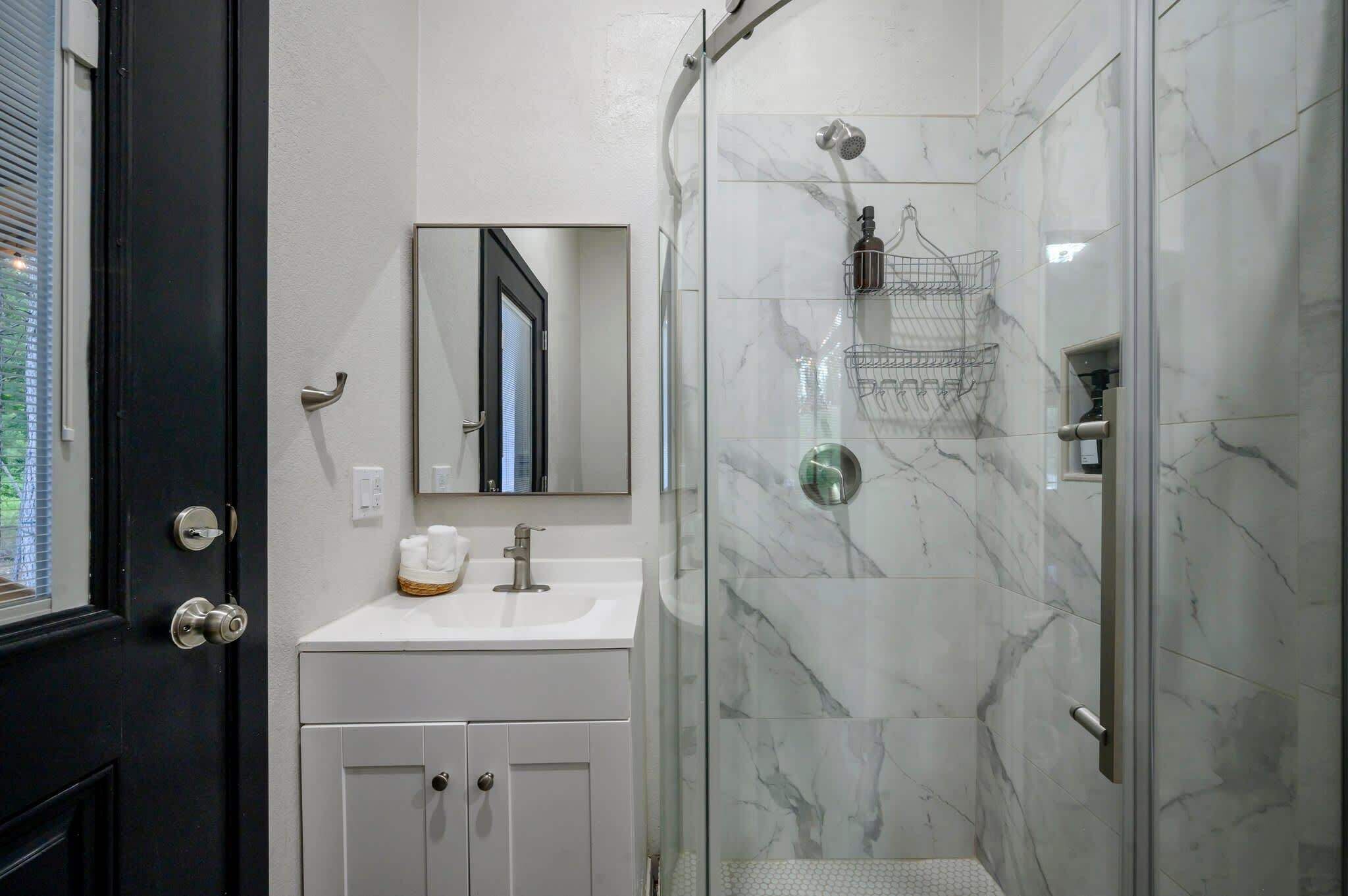A bathroom with a white vanity, a mirror, a shower with a glass door, and a marble-tiled wall.