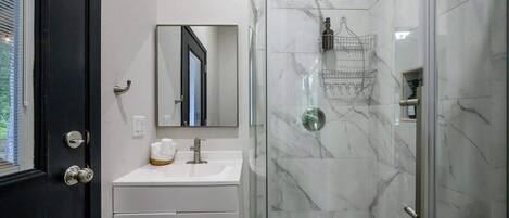 A bathroom with a white vanity, a mirror, a shower with a glass door, and a marble-tiled wall.