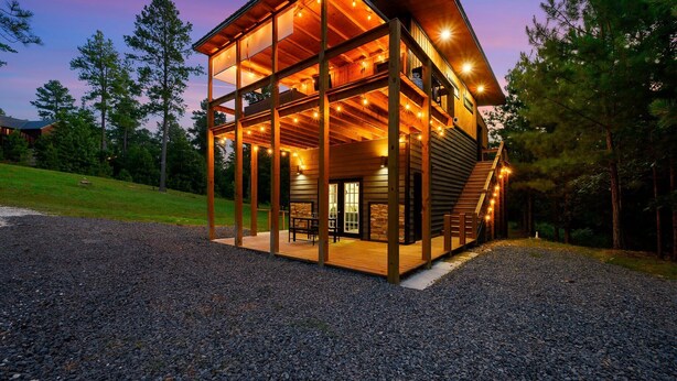 A large, multi-level wooden cabin with a spacious deck and multiple windows, surrounded by a gravel driveway and trees. - Double Magic - Unique Cabin for 6 - Pond, Hot Tub (Broken Bow)