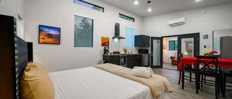A modern hotel room with a bed, a dining table with chairs, a kitchenette, and a ceiling fan.