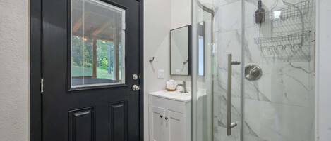 Un baño moderno con puerta negra, cabina de ducha de cristal, lavabo blanco y espejo sobre él.
