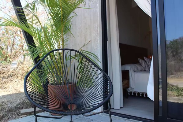Une chaise avec un pot de plante, un lit avec une literie blanche et une vue sur l’extérieur.
