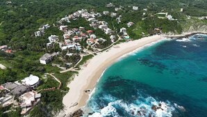 An aerial view of a coastal area with a beach, houses, and a resort with a pool.