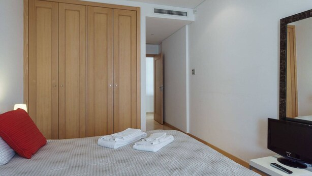 A bedroom with a bed, a TV, a TV stand, a red pillow, and a wardrobe. - Vilamourasun Aquamar 112 (Loulé)