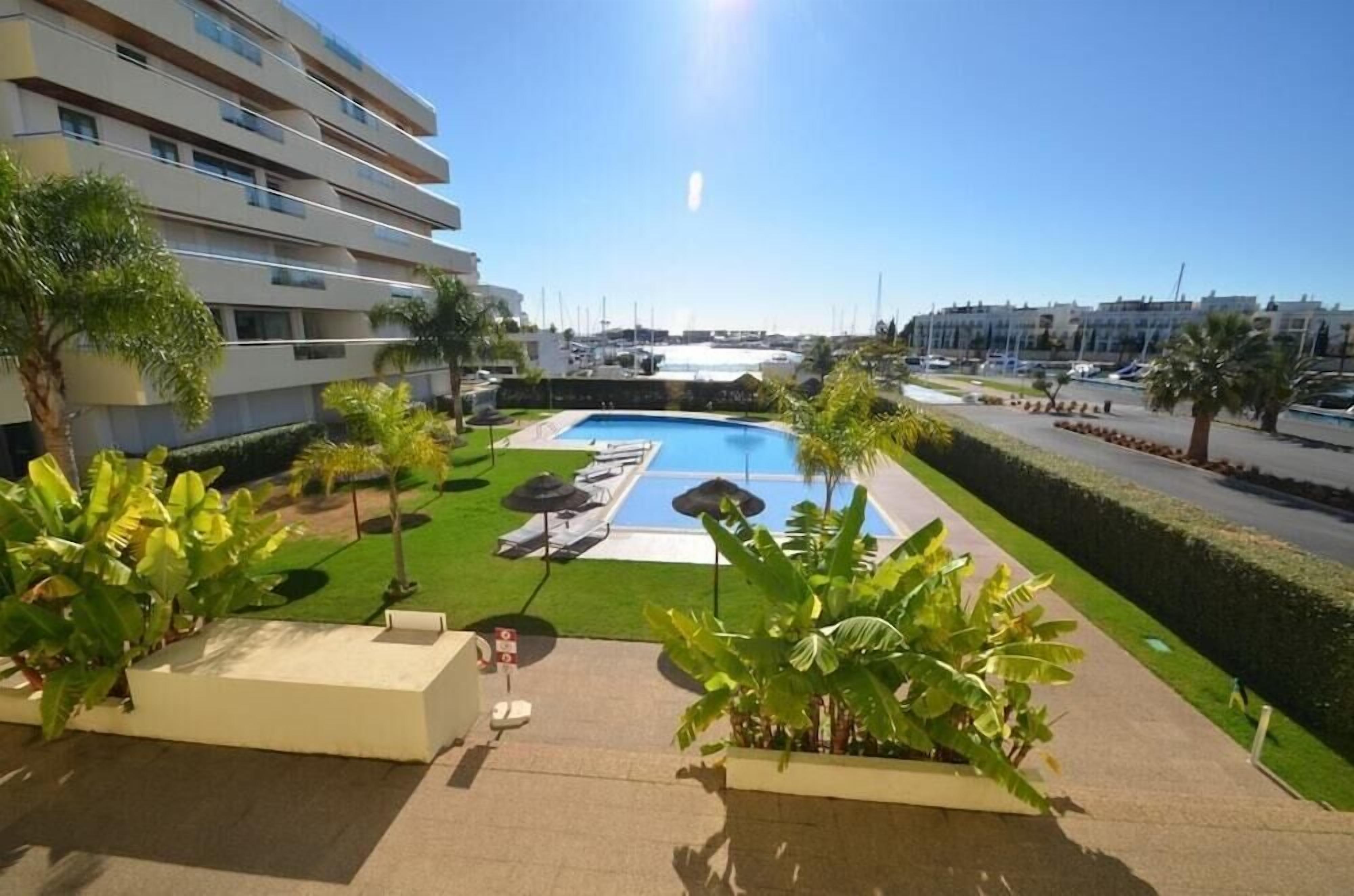 A pool area with lounge chairs and umbrellas, surrounded by greenery and a clear sky.
