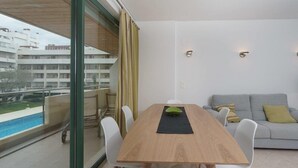 A modern living room with a wooden dining table, white chairs, a grey sofa, and a view of a swimming pool and buildings. - Vilamourasun Aquamar 112 (Loulé)