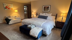 Un dormitorio con una cama grande, dos mesitas de noche, un pouf y un cuadro decorativo en la pared.