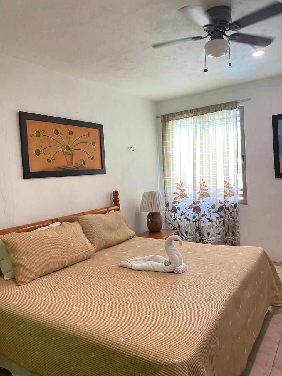 A bedroom with a bed, a ceiling fan, a window with curtains, a lamp, and a framed picture on the wall.