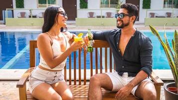 A couple sitting by a poolside with drinks, a wooden bench, and a potted plant.