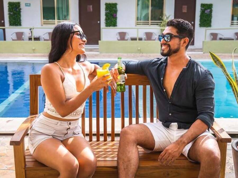 A couple sitting by a poolside with drinks, a wooden bench, and a potted plant.