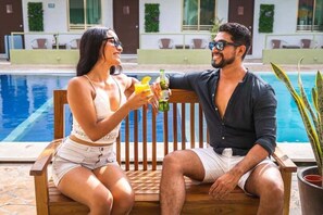 A couple sitting by a poolside with drinks, a wooden bench, and a potted plant. - Pelican Inn at Las Dunas Resort (Progreso)