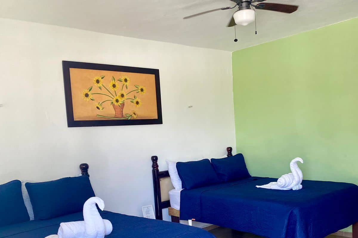 Two single beds with blue bedding, a ceiling fan, and a framed painting of sunflowers on the wall.
