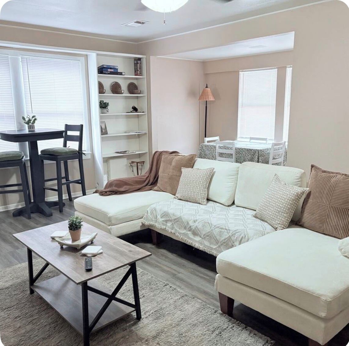 A living room with a sectional sofa, a coffee table, a bookshelf, and a dining area with chairs and a table.