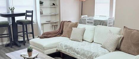 A living room with a sectional sofa, a coffee table, a bookshelf, and a dining area with chairs and a table.