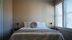 Ein ordentlich bezogenes Bett mit einer beigen Decke und zwei Kissen, ein Nachttisch mit einer Lampe und ein Fenster mit Jalousien.
