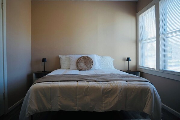 A neatly made bed with a beige blanket and two pillows, a bedside table with a lamp, and a window with blinds.