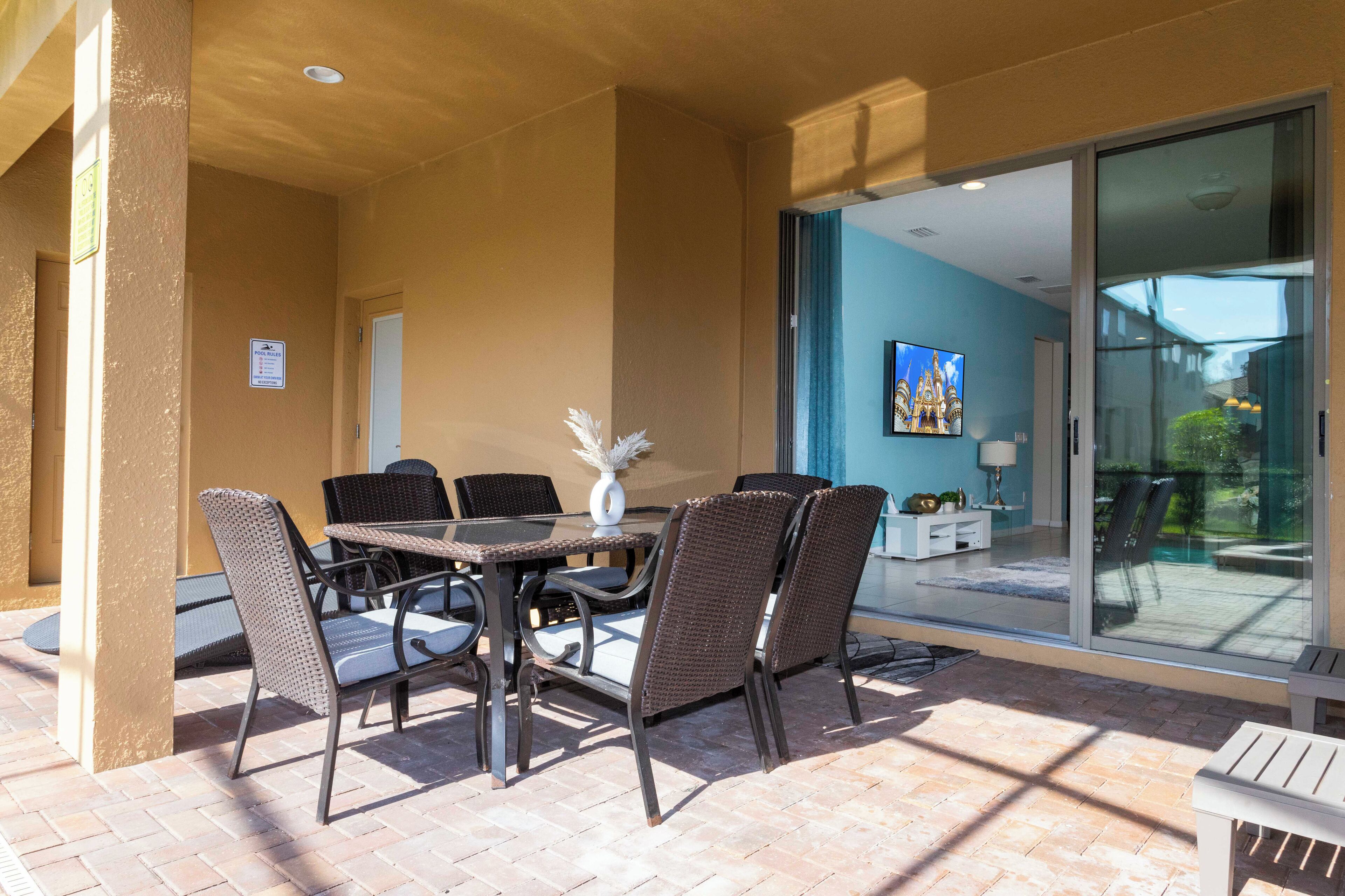 Un patio al aire libre con una mesa y sillas, un banco, y un ventanal de vidrio que da a una habitación con pared azul y un cuadro.